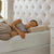 A woman with curly hair, wearing a gray sleeveless top and beige pants, lies on her side on a bed, smiling peacefully as she rests her head on the La-Z-Boy Hybrid Bliss Memory Foam Pillow in a cozy, well-lit bedroom.