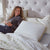 A woman with curly hair, in a gray cardigan and pants, sits up in bed against a tufted white headboard, smiling and holding a cup. The La-Z-Boy Hybrid Bliss Memory Foam Pillow adds comfort to the cozy, inviting scene.
