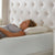 A woman with curly hair relaxes on a white mattress, her head resting on the La-Z-Boy Hybrid Bliss Memory Foam Pillow with a moisture-wicking cover. She wears a sleeveless grey top, near a light tufted headboard and white nightstand.