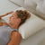 A woman with curly hair lies comfortably on her back, eyes closed, resting on a La-Z-Boy Hybrid Bliss Memory Foam Pillow. She wears a gray sleeveless top and bracelet, relaxing on a light-colored, textured bed surface.