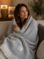 A woman sits on a sofa wrapped in a soft, gray herringbone 100% Organic Cotton Algarve Throw Blanket from Delilah Home, its fringe draped gently. She smiles softly, gazing off to the side. Behind her, a lamp, candle, framed photo, and leafy plant add to the cozy and inviting atmosphere.