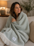 A woman with wavy brown hair sits on a beige couch, wrapped in a soft, light green Delilah Home 100% Organic Cotton Algarve Throw Blanket featuring a herringbone pattern and white fringed edges. She smiles gently, with a lamp, framed art, and a vase of greenery in the softly-lit background.