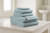 A neatly folded stack of Delilah Home’s 100% Organic Cotton Bath Towels Collection in soft light blue rests on a marble countertop, with a blurred sink and window in the background for a calm, airy bathroom feel.