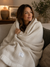 A woman sits on a couch, wrapped in a Delilah Home 100% Organic Cotton Algarve Throw Blanket in cream. She smiles softly and gazes to the side. The cozy living room is warmly lit, with a lamp, artwork, and a vase of greenery resting on a wooden table in the background.