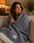 A woman sits on a beige couch, wrapped in a Delilah Home 100% Organic Cotton Algarve Throw Blanket featuring a blue and white herringbone pattern with fringed edges. She smiles softly while looking to the side, surrounded by cozy decor, a lamp, and a plant in the warmly lit background.