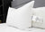 A Pillow Factory Downlite 50/50 Waterfowl Feather/Grey Down Pillow with a care tag rests in front of another medium support pillow on a neatly made bed with white sheets and a gray headboard, shelving partially visible in the modern room.