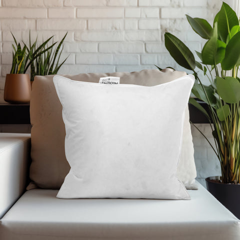 A comfy white Pillowtex throw pillow with a Pillowtex white duck feather & down pillow insert and a visible tag sits on a sofa, accessorized by green potted plants and a modern white brick backdrop, creating a tranquil and stylish home.