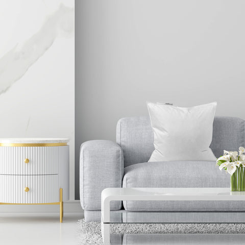 A minimalist modern living room scene featuring a grey sofa with a Pillowtex Pillow Insert | White Duck Feather & Down cushion, a white and gold cabinet, and a vase of fresh flowers, set against a clean, white wall.