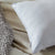 A close-up of a white La-Z-Boy Fiber Density Pillow with a branded tag rests on beige sheets beside a chunky gray knit blanket, creating a cozy, inviting bed with soft textures and neutral tones.