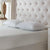 A neatly made bed displays two white La-Z-Boy Fiber Density Pillows side by side on a textured white mattress, with a large, tufted beige headboard and a partially visible bedside table holding a wooden lamp and decorative object.
