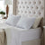 A neatly made bed with a tufted beige headboard features two white pillows, including the plush La-Z-Boy Fiber Density Pillow, and a cream quilted pillow. White sheets and a light beige comforter complete the look beside a nightstand with a lamp, vase, and potted plant.
