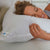 A woman with curly hair smiles while sleeping on her side, hugging a fluffy white La-Z-Boy Fiber Density Pillow. She rests on a quilted white mattress with a white headboard and nightstand visible in the background.