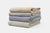Four American Blossom Linens Herringbone Weave Blankets, crafted from organic cotton in the USA, are stacked in beige, light gray, blue, and light green. Their subtle pattern and heirloom quality add cozy style to any home.
