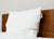 A neatly made bed with a wooden headboard features two stacked Pillowtex White Duck Down & Feather Pillows (75% feather/25% down) by Pillowtex, each in white with a visible care label, against a plain light beige wall.