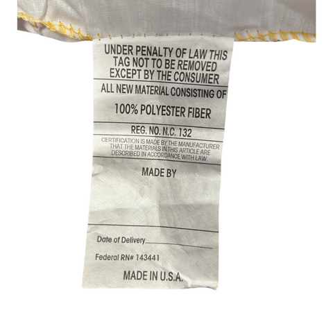A close-up of a crumpled white care label attached to a JS Fiber Gold Choice Polyester Pillow Featured in Some Embassy Suites | Extra Loft, displaying black text that includes legal warnings, a certification number, and "made in u.s.a.