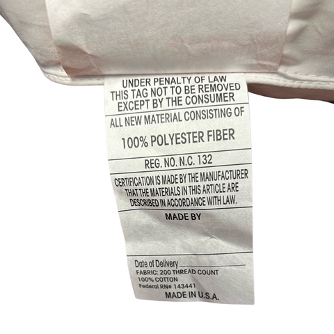 A close-up of a white care label attached to a JS Fiber Invista Comforel Gussetted Pillow | Medium-Firm displaying text. It includes washing instructions, fabric content as "100% polyester," and other regulatory information. The label states