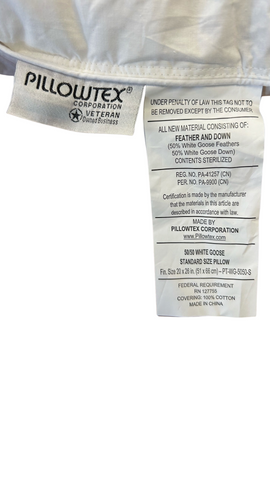 A close-up of a Pillowtex High End White Goose Down Pillow label showing material information, care instructions, and a warning about removal of the tag, sewn on a luxury Pillowtex pillow under a soft-focused background.