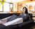 A woman in gray loungewear relaxes on a black sofa, legs elevated on the Sleep Again Pillows Extra-Wide 30” Wedge Pillow for acid reflux relief. She holds a remote in a bright, modern living room with wooden beams and fresh flowers.