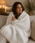 A woman with wavy brown hair sits on a beige couch, wrapped in a cozy Delilah Home 100% Organic Cotton Algarve Throw Blanket in cream, featuring sustainable luxury, a fringe detail, and a small label. She smiles gently while looking to the side, surrounded by the warm glow of a softly lit room decorated with a lamp, candle, framed photo, and plant.