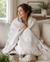 A woman sits on a light sofa, wrapped in a cream-colored 100% Organic Cotton Algarve Throw Blanket by Delilah Home, holding it close and smiling. Her wavy brown hair is pulled back and she wears a cozy sweater. A mug rests on the table in the foreground, while soft home decor fills the background, creating an atmosphere of sustainable luxury and comfort.