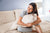 A woman in light gray pajamas sits cross-legged on a bed, smiling and hugging a beige pillow covered with American Blossom Linens’ Cotton Bed Pillowcases with Piping, made from 100% USA cotton. Natural light brightens the cozy, neutral room.