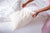 A person in pink pajamas gently slides a white pillowcase onto the American Blossom Linens American Made Wool Filled Pillow—organic cotton, adjustable firmness—on an unmade white bed, creating a bright and clean scene.