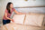 A woman with long brown hair and a pink shirt smiles as she makes the bed, arranging beige American Blossom Linens Colored Cotton Bed Pillowcases—crafted from natural heirloom cotton, made in the USA—on a wooden headboard.