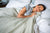 A dark-haired woman lies on her side in bed, smiling with her eyes closed as she cuddles the American Blossom Linens Herringbone Weave Blanket, made from organic cotton in the USA. The peaceful room features a cozy, inviting atmosphere.