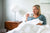 A blonde woman sits up in bed, smiling with a white "THANKFUL" mug. She relaxes under a comforter with American Blossom Linens Cotton Bed Pillowcases with Piping, made from 100% USA cotton, beside a modern lamp and wooden nightstand.