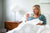 A woman with blonde hair sits up in bed under a white comforter, smiling while holding a "THANKFUL" mug. Her head rests on American Blossom Linens Cotton Bed Pillowcases with Piping, 100% USA cotton and American made. The room is bright and cozy.