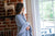 A woman with wavy, shoulder-length hair stands indoors by a brick wall, wrapped in the American Blossom Linens Herringbone Weave Blanket, made from organic cotton in the USA, gazing out a sunlit window.