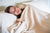 A woman with gray hair sleeps peacefully, covered to her shoulders with the Herringbone Weave Blanket by American Blossom Linens. Made in the USA from organic cotton, this heirloom-quality blanket softly illuminates her serene rest on white bedding.