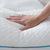 A hand gently presses down on the white Malouf FlexFill™ + HyperChill™ Pillow, showcasing its softness and cushioning. The pillow features a textured, quilted surface with diamond and circular patterns. The blue trim along the edge complements its HyperChill™ Cooling Cover. The scene suggests comfort and quality.