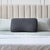 A black Malouf HexCurve™ Pillow with a hexagonal pattern stands upright against a beige tufted headboard on a neatly made bed with light blue sheets, white quilt, and two light blue pillows behind the cooling Malouf pillow.