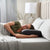 A woman with brown hair in a green shirt lies on her side in bed, smiling with eyes closed as she rests on the Malouf HexCurve™ Pillow, known for its ergonomic and cooling comfort. Soft sheets cover the bed, with a lamp and vase nearby.
