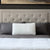 A bed with a tufted beige headboard features two dark gray pillows and a Malouf Quilted Convolution™ Pillow by Malouf in front. The bed is neatly made with a dark gray sheet, white blanket, and has abstract artwork above the headboard.
