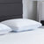 Two Carpenter Comfort Tech Dual Comfort Foam Pillows, featuring textured covers and light blue trim, rest on a white quilted bed with a dark gray upholstered headboard and a white lamp, creating a clean, minimalistic look.