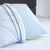 A Comfort Tech Dual Comfort Foam Pillow by Carpenter in light blue with perforations is propped on a bed with a white and pale blue striped cover and matching pillowcase, with a gray upholstered headboard visible in the background.