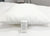 A Pillow Factory Comforel Pillow, featured at Sheraton Hotels, with subtle stripes and a hypoallergenic white cover rests on a quilted bed. A tag with care instructions is attached; blurred bedding and furniture appear in the background.