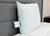 A medium, white JS Fiber Hollofil II Pillow stands upright on a white mattress against a gray upholstered headboard. A tag with text and a small blue logo is visible on the side seam. A small wooden nightstand appears in the background.