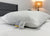 The Manchester Mills Envirosleep Gold Plus Pillow with firm polyester fiber fill, featuring visible tags, rests on a neatly made bed with white bedding and a gray headboard. A lit beige lamp on a round table enhances the serene setting.
