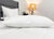 A close-up of a neatly made bed features the Pillowtex Conformance Pillow | Soft Support by Pillowtex on a white comforter, with a grey upholstered headboard, potted plant, and lit floor lamp with a beige shade in the background.