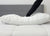 Wearing black pants, a person presses down on the Envirosleep Gold Plus Pillow by Manchester Mills, its firm polyester fiber fill compressing atop a white quilted bed with a grey upholstered headboard in the background.