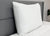 An Envirosleep Dream Support Pillow by Manchester Mills is propped vertically against a gray upholstered headboard on a neatly made bed with a white comforter, complemented by a potted plant in the minimal, clean bedroom setting.
