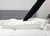 A person presses their arm into the Down Etc. Fairfax Pillow—featured at St. Julien Hotel & Spa®—on a white bed with a grey upholstered headboard; tags hang from the pillow, and a plant is partly visible in the background.