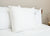 A single large white Keeco Best Western® Aurora Pillow | Medium Polyester Pillow stands upright on a neatly made bed with white bedding. Two more pillows are partially visible behind it, with a tag on the front pillow.
