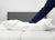 A person in navy blue pajamas presses their arm into a Down Etc. 50% White Goose Down / 50% White Goose Feather Pillow on a white mattress with a gray upholstered headboard and plain white background; the pillow’s side has a small tag.