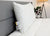 A clean, white Manchester Mills Envirosleep Gold Plus Pillow | Firm Polyester Fiber Fill with attached tags rests against a gray headboard on a neatly made bed with white comforter; a potted plant and lamp complete the softly lit, hotel-quality scene.