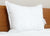 The Pacific Coast® Natural Feather Pillow by Pacific Coast Feather Company, with its tag visible, rests against a wooden headboard on white bedding, set against a neutral wall for a clean, simple bedroom look.