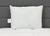 A plain white JS Fiber Hollofil II Pillow | Medium with a small left tag stands upright on a white quilted surface. The dark gray, paneled headboard enhances the clean, modern look of this hypoallergenic pillow by JS Fiber.